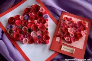 Na zdjęciu znajdują się dwie kartki papieru w kolorze czerwonym z naklejonymi kawałkami papieru zwiniętymi w ruloniki różnej wielkości. Ruloniki ułożone są w kształcie serca. Na jednej z kartek, przyklejony jest mały prostokątny kawałek białego papieru z napisem „kocham Cię”. Kartki leżą na jasno fioletowym materiale.
